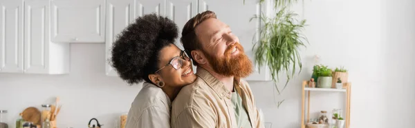 Alegre mujer afroamericana en gafas de pie cerca de novio en la cocina, pancarta - foto de stock