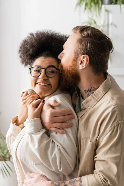 Бородатий чоловік цілує і обіймає усміхнену афроамериканську дівчину вдома — стокове фото