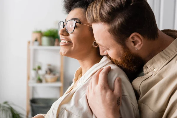 Bearded uomo baciare positivo africano americano fidanzata in occhiali da vista a casa — Foto stock
