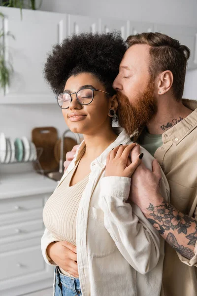 Tatuado homem abraçando e beijando sorridente afro-americano namorada em casa — Fotografia de Stock