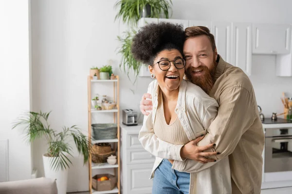 Alegre africana americana mujer abrazando novio y mirando a la cámara en casa - foto de stock