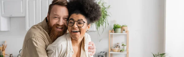 Positive bearded man hugging cheerful african american girlfriend in kitchen, banner — Stock Photo
