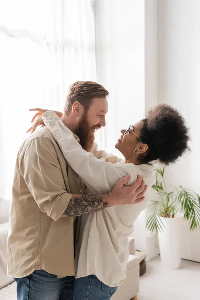 Side view of bearded man hugging and looking at happy african american girlfriend at home — Stock Photo