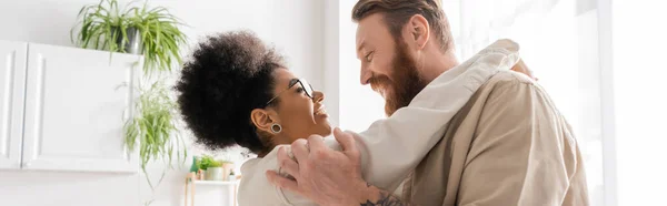 Side view of positive african american woman hugging bearded boyfriend at home, banner — Stock Photo