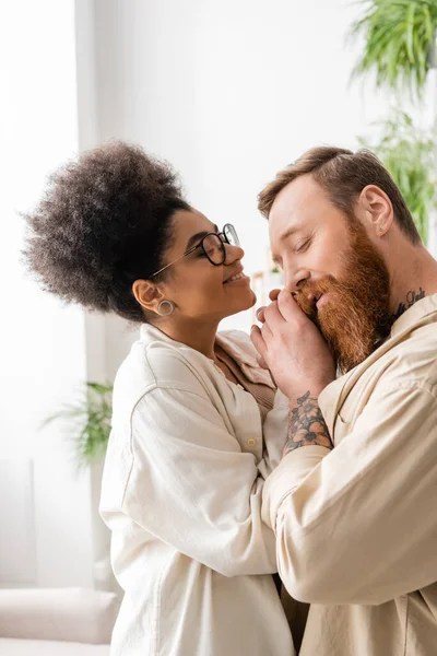 Hombre barbudo tocando la mano de la sonriente novia afroamericana en casa - foto de stock