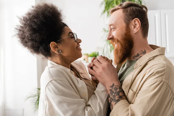 Vista lateral do homem tatuado segurando a mão de uma namorada afro-americana muito feliz em casa — Fotografia de Stock
