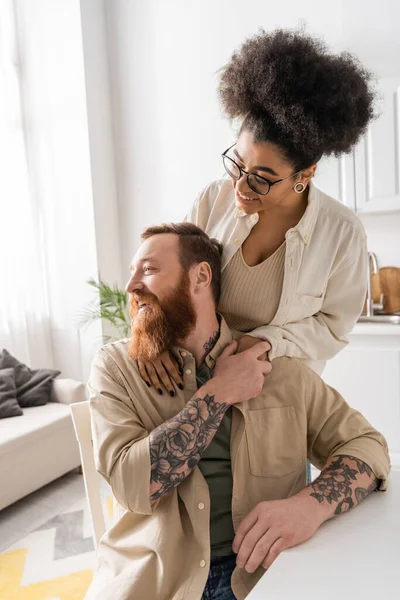 Positive african american woman hugging tattooed boyfriend in kitchen — Stock Photo