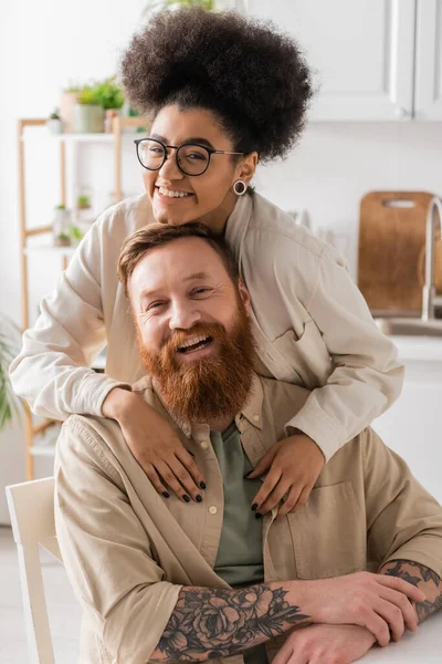 Sorridente donna africana americana che abbraccia il fidanzato barbuto e guarda la fotocamera a casa — Foto stock