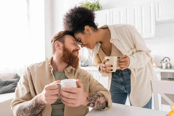 Side view of smiling african american woman holding cup near tattooed boyfriend in kitchen — Stock Photo