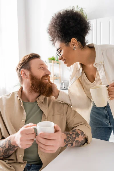 Smiling african american woman with cup of coffee touching bearded man at home — Stock Photo