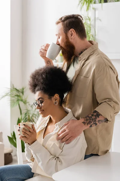 Tatuado homem beber café perto satisfeito afro-americano namorada em casa — Fotografia de Stock
