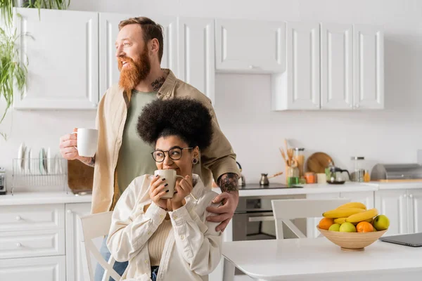 Positive bearded man holding cup of coffee and hugging african american girlfriend in kitchen — Stock Photo