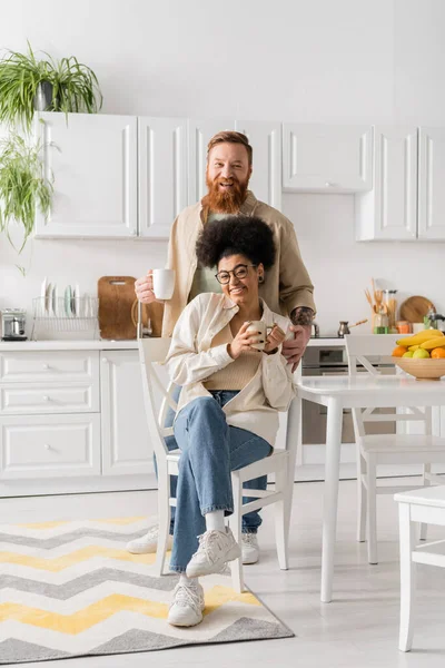 Alegre pareja multiétnica sosteniendo tazas de café y mirando a la cámara en la cocina - foto de stock