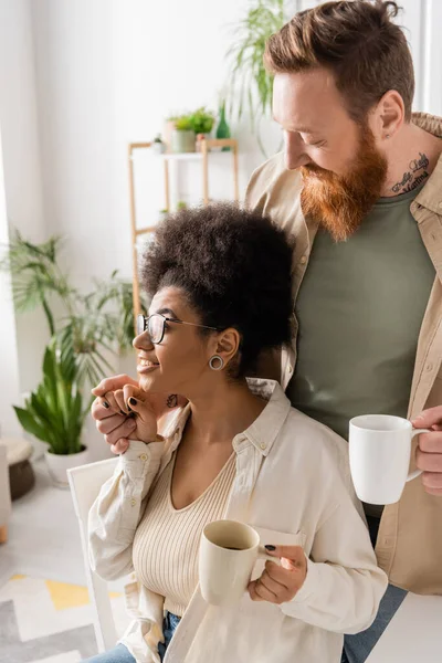 Bearded man holding cup and hand of smiling african american girlfriend at home — Stock Photo