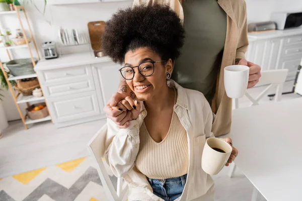Smiling african american woman holding hand of boyfriend and cup of coffee in kitchen — Stock Photo