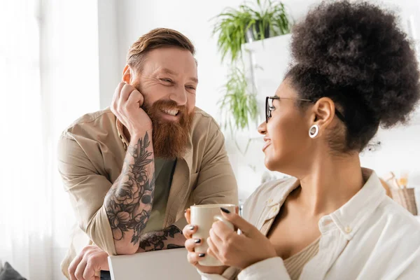 Smiling tattooed man looking at african american girlfriend with cup of coffee in kitchen — Stock Photo