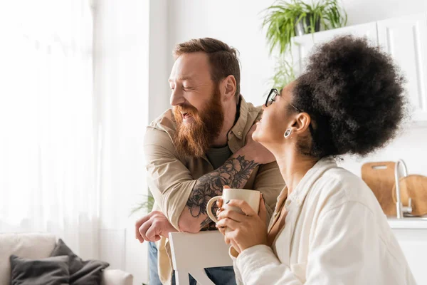 Überglückliche Afroamerikanerin hält Kaffeetasse neben bärtigen Freund zu Hause — Stockfoto