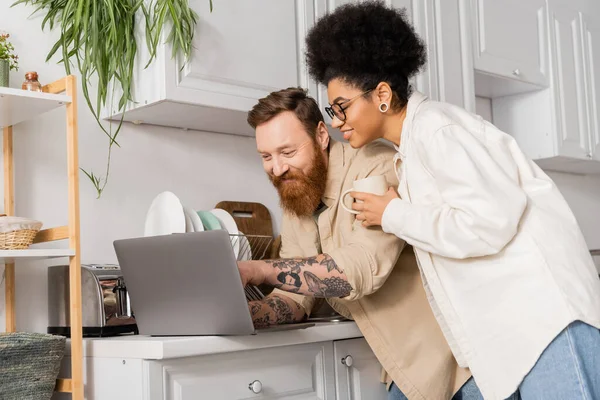 Homme tatoué souriant utilisant un ordinateur portable près de petite amie afro-américaine avec café dans la cuisine — Photo de stock
