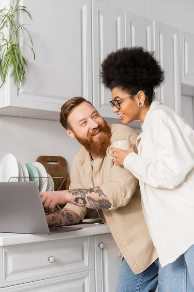 Homme tatoué utilisant un ordinateur portable et regardant la petite amie afro-américaine avec du café dans la cuisine — Photo de stock