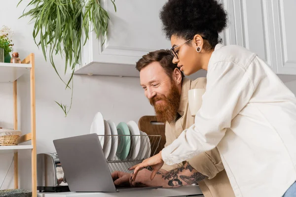 African american woman with cup pointing at laptop near bearded boyfriend in kitchen — Stock Photo