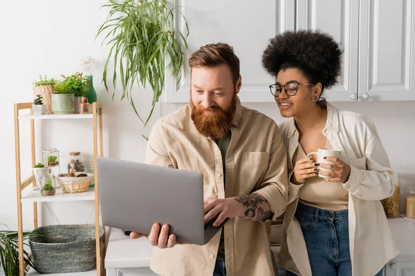 Tattooed man using laptop near african american girlfriend with cup in kitchen — Stock Photo