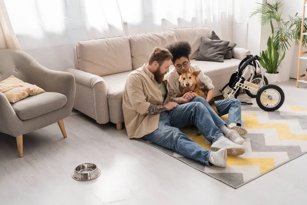 Tätowierter Mann sitzt neben afroamerikanischer Freundin und behindertem Hund im Wohnzimmer — Stockfoto