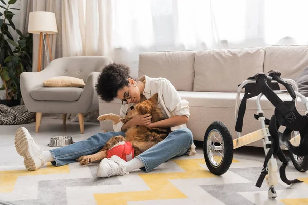 Joven mujer afroamericana abrazando perro discapacitado cerca de silla de ruedas en sala de estar - foto de stock
