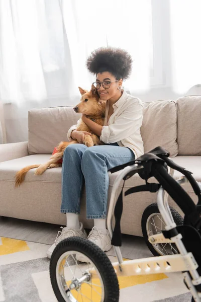 Sonriente mujer afroamericana abrazando a un perro discapacitado cerca de una silla de ruedas borrosa en la sala de estar - foto de stock