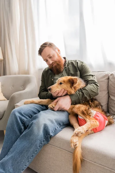 Uomo barbuto abbracciare cane handicappato mentre seduto sul divano a casa — Foto stock