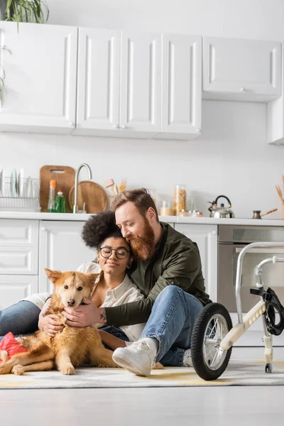Multiethnic couple sitting near disabled dog on carpet near wheelchair in kitchen — Stock Photo