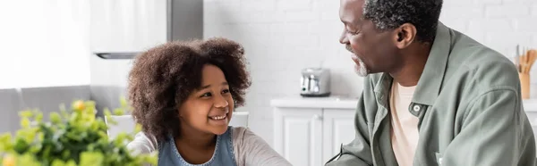 Happy african american grandfather looking at curly granddaughter in kitchen, banner — Stock Photo