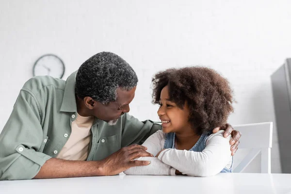 Feliz avô afro-americano abraçando a neta encaracolada na cozinha — Fotografia de Stock