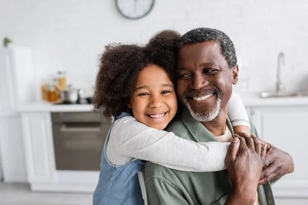 Portrait of happy african american girl with curly hair smiling and embracing cheerful grandfather at home — Stock Photo