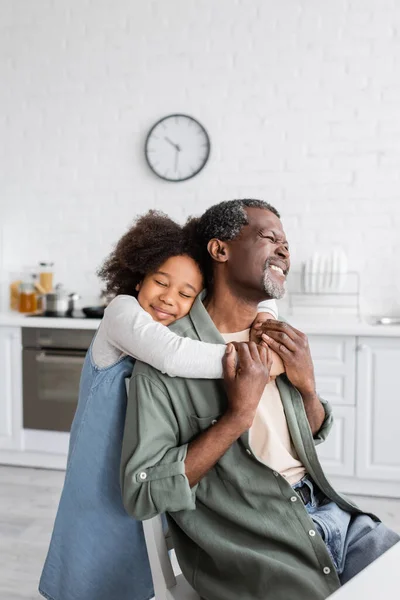 Happy african american girl with curly hair smiling and embracing pleased grandfather at home — Stock Photo