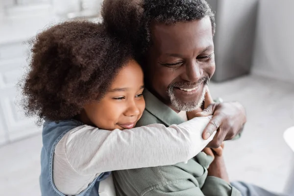 Menina americana africana feliz com cabelo encaracolado abraçando avô alegre em casa — Fotografia de Stock