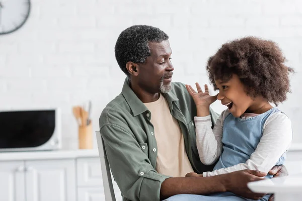 Étonnante fille afro-américaine avec des cheveux bouclés gesticulant tout en parlant avec grand-père joyeux à la maison — Photo de stock