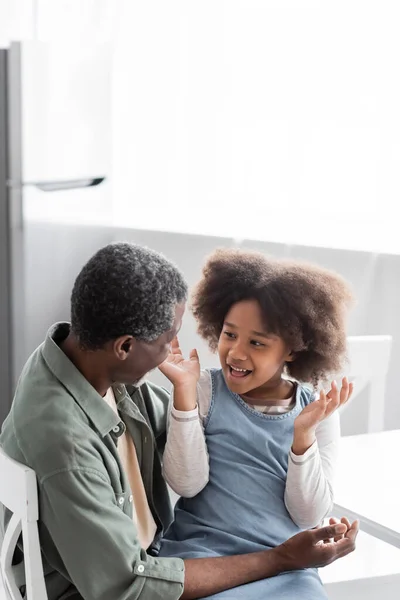 Aufgeregtes afrikanisch-amerikanisches Mädchen mit lockigem Haar gestikuliert beim Gespräch mit dem fröhlichen Großvater zu Hause — Stockfoto