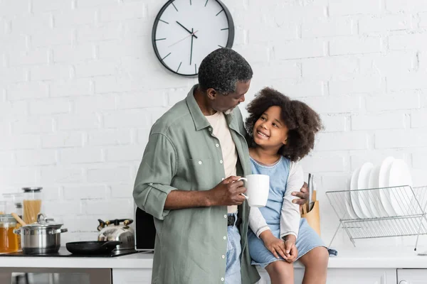 Bearded african american grandfather holding cup of coffee and hugging happy child in kitchen — Stock Photo