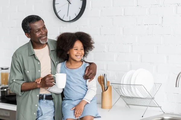 Bärtiger afrikanisch-amerikanischer Großvater hält Tasse mit Kaffee und umarmt freudiges Kind in der Küche — Stockfoto