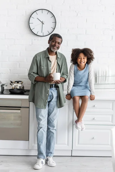 Grand-père afro-américain d'âge moyen tenant tasse avec café près enfant joyeux assis sur le plan de travail de la cuisine — Photo de stock