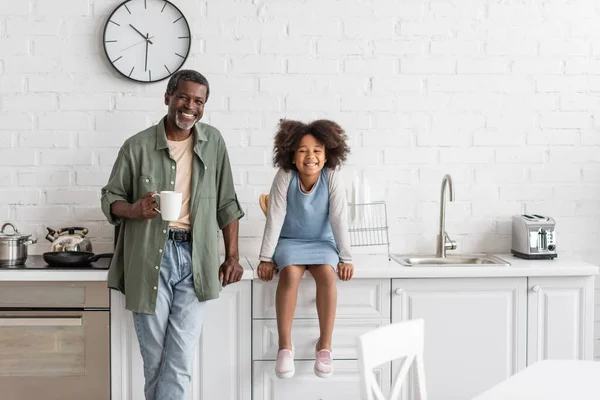 Hombre afroamericano de mediana edad sosteniendo taza con café cerca de la nieta feliz sentado en encimera de la cocina - foto de stock