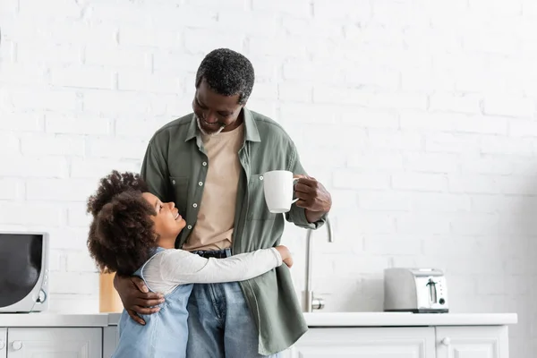 Happy african american kid hugging grandfather holding cup with coffee in kitchen — Stock Photo