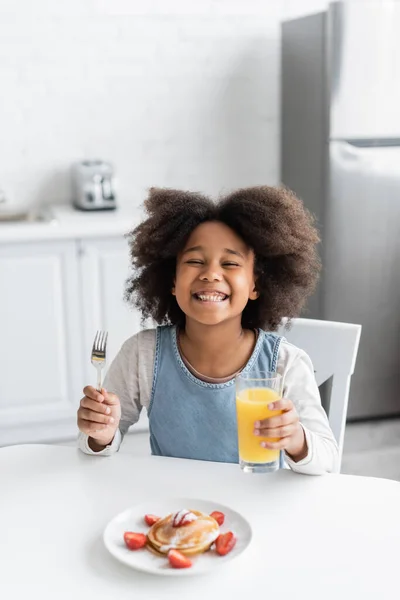 Chica afroamericana feliz sosteniendo tenedor y vaso de jugo de naranja cerca de panqueques mientras disfruta del desayuno - foto de stock