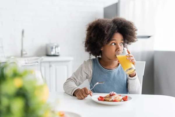 Rizado afroamericano chica bebiendo jugo de naranja y sosteniendo tenedor cerca de panqueques mientras desayuna - foto de stock