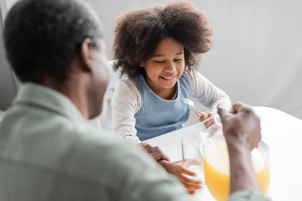 Happy african american girl holding fork while grandpa pouring orange juice during breakfast — Stock Photo