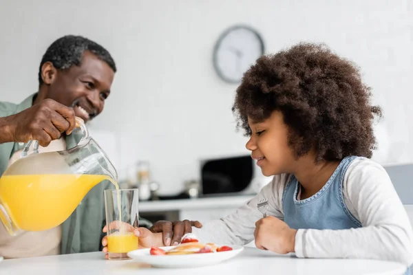 Счастливая африканская американская девушка смотрит на стекло, а дедушка наливает апельсиновый сок во время завтрака — стоковое фото