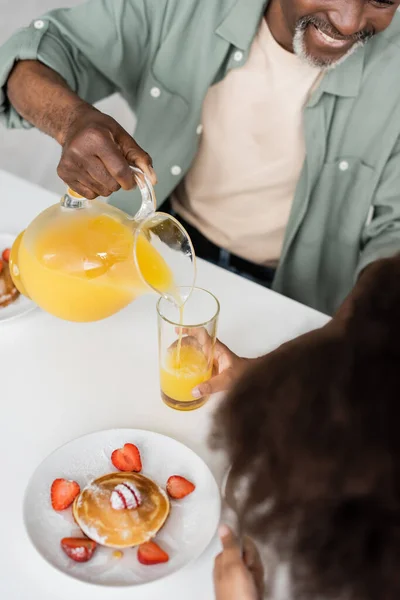 Vue du dessus de l'homme afro-américain heureux versant du jus d'orange près de la petite-fille bouclée pendant le petit déjeuner — Photo de stock