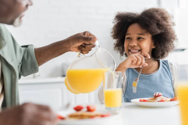 Щаслива афроамериканська дівчина, дивлячись на діда, що поливає апельсиновий сік під час сніданку — стокове фото