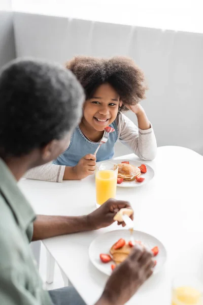 Щаслива афроамериканська дівчина дивиться на діда, що їсть млинці під час сніданку — стокове фото