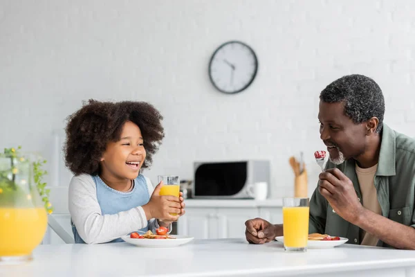 Menina americana africana feliz segurando vidro de suco de laranja enquanto toma café da manhã com o avô — Fotografia de Stock
